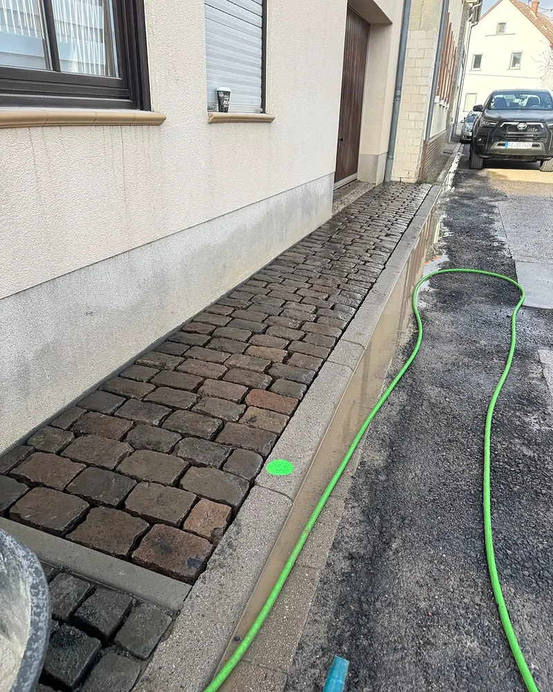 Detailaufnahme des frisch verlegten und eingeschlämmten Naturstein-Kopfsteinpflasters entlang der Hauswand in Mainz