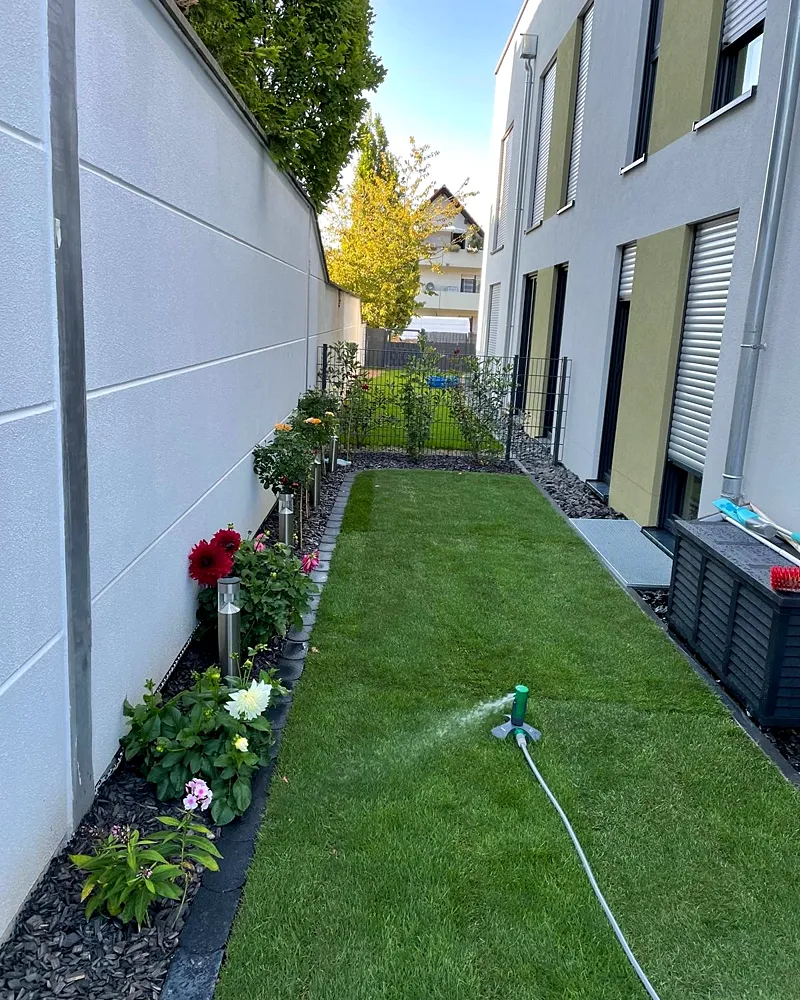 Schmaler Gartenstreifen mit frischem Rollrasen, Blumenbeet und Sprinkler
